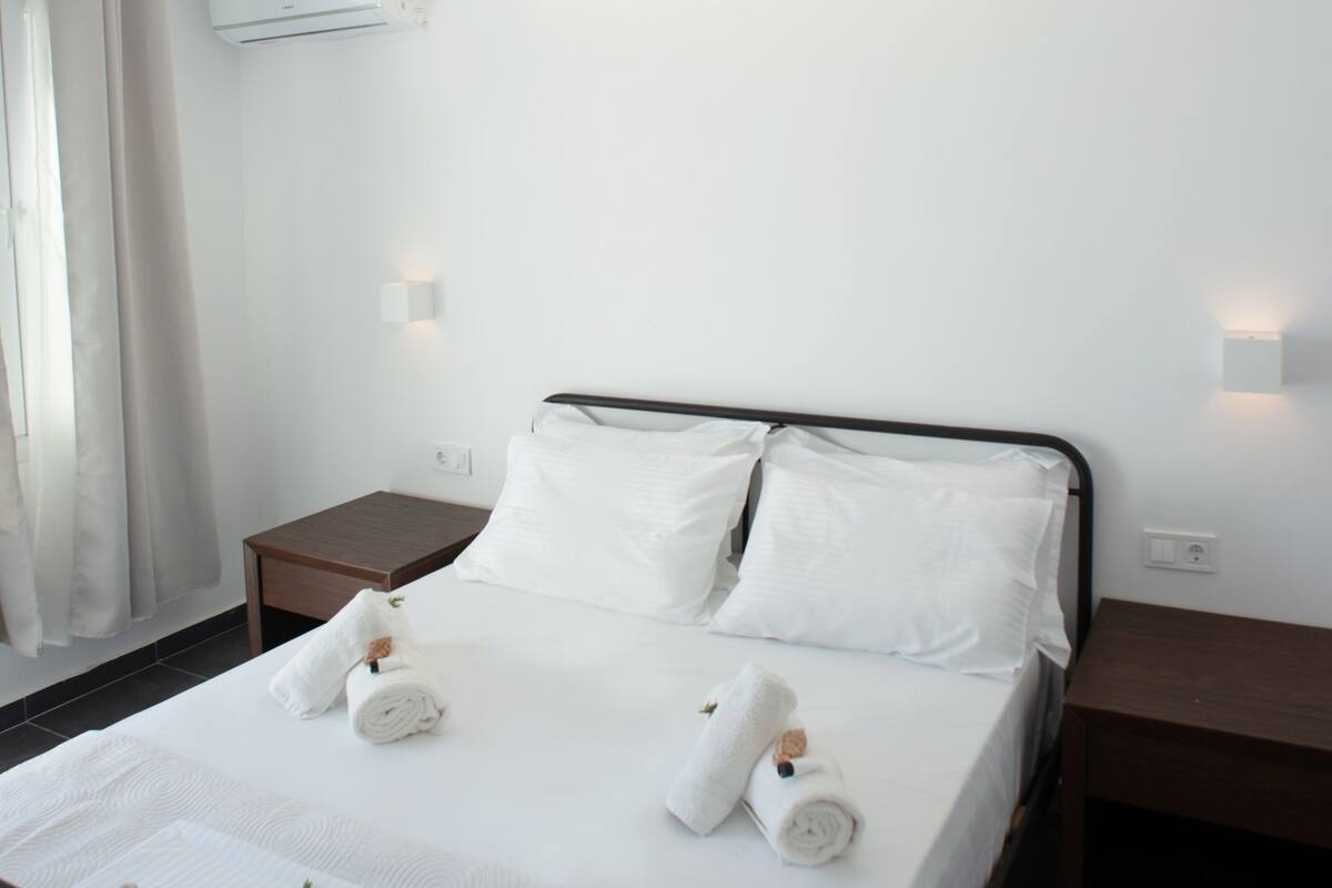 Een net opgemaakt bed met witte lakens en handdoeken, een nachtkastje, een wandlamp en een airconditioner aan de muur.