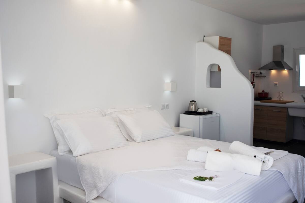 Een moderne hotelkamer met een net opgemaakt bed, een klein nachtkastje, een geïntegreerd keukenhoekje met spoelbak en kookplaat, en een groot raam dat natuurlijk licht biedt.