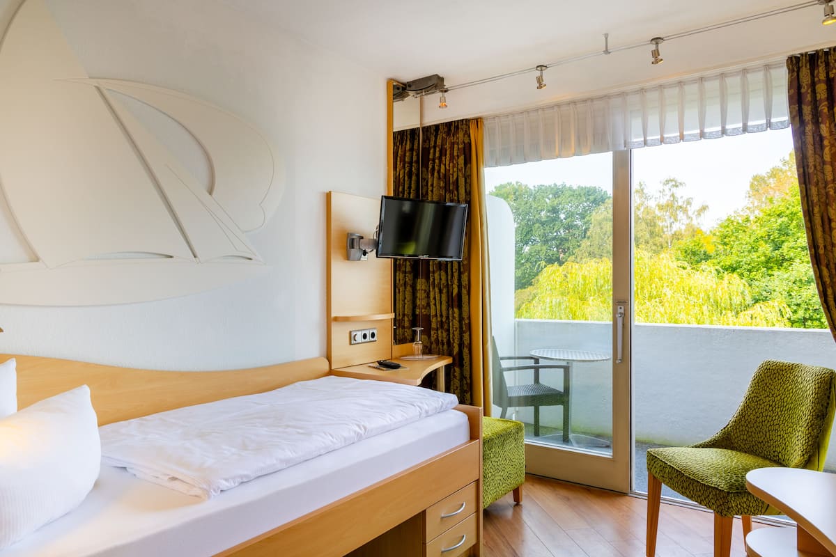 Ein Hotelzimmer mit Bett, Schreibtisch mit Fernseher, Stuhl und Balkon mit Blick auf Grünflächen.