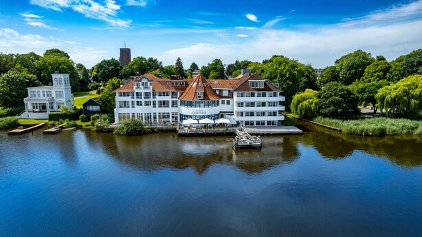 A large, multi-story hotel with a red-tiled roof, situated by a calm lake with docked boats. - Seehotel Fährhaus (Bad Zwischenahn)