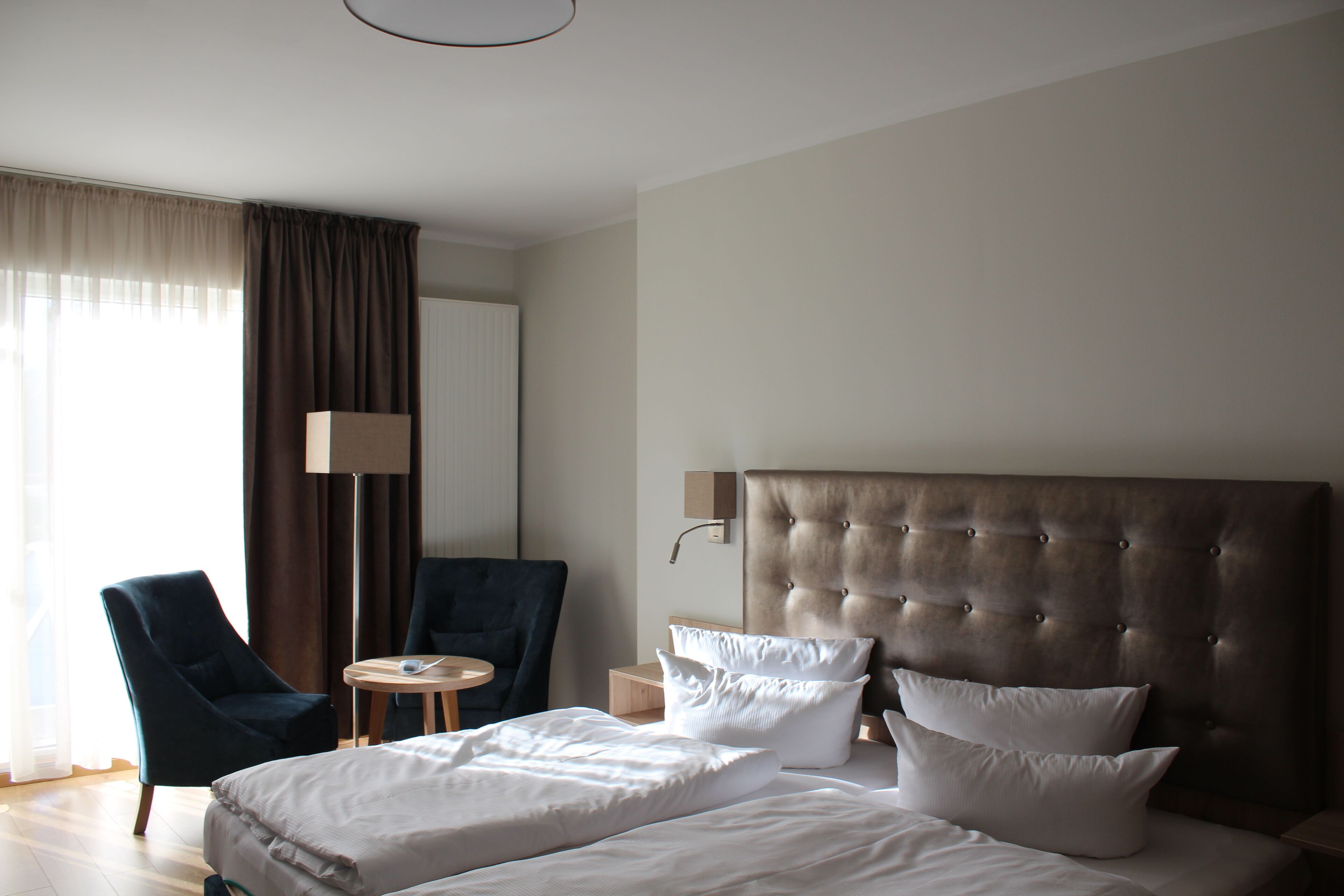 Ett hotellrum med en stor säng, två kuddar, ett sängbord, en lampa, en stol och ett fönster med gardiner.