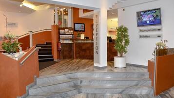 Área de recepção de hotel com escadaria de mármore, balcão de recepção e uma tela de TV exibindo um mapa.
