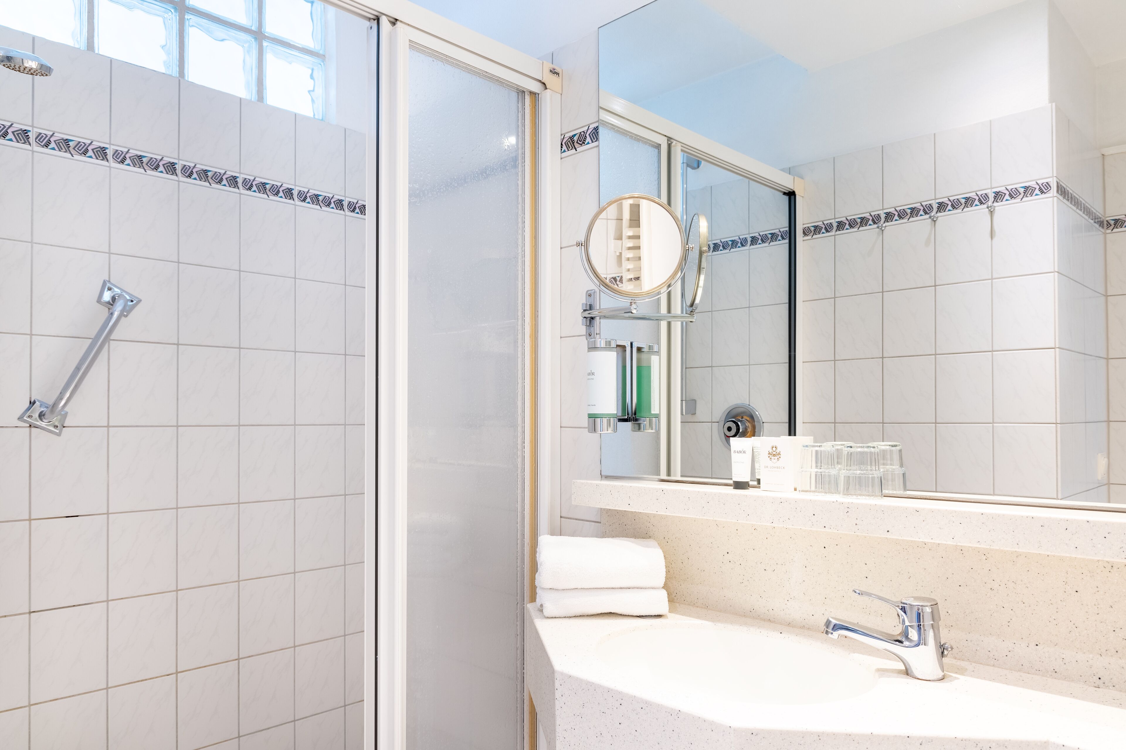 A bathroom with white tiled walls, a shower with a glass door, a sink with a mirror, and a towel rack.