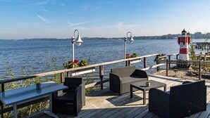 A deck with outdoor seating, a lighthouse, and a view of a calm body of water. - Seehotel Fährhaus (Bad Zwischenahn)
