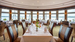 A dining area with round tables, chairs, and a view of the water. - Seehotel Fährhaus (Bad Zwischenahn)