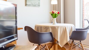 Ein Essbereich mit einem Tisch, der mit einer gestreiften Tischdecke bedeckt ist, zwei schwarzen Stühlen, einer Vase mit Tulpen, einem Fernseher und einem Bild an der Wand.