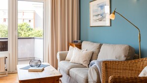 Ein Wohnzimmer mit einer Couch, einem Korbsessel, einem Tisch mit einem Buch, einer Lampe und einem Gemälde an der Wand.