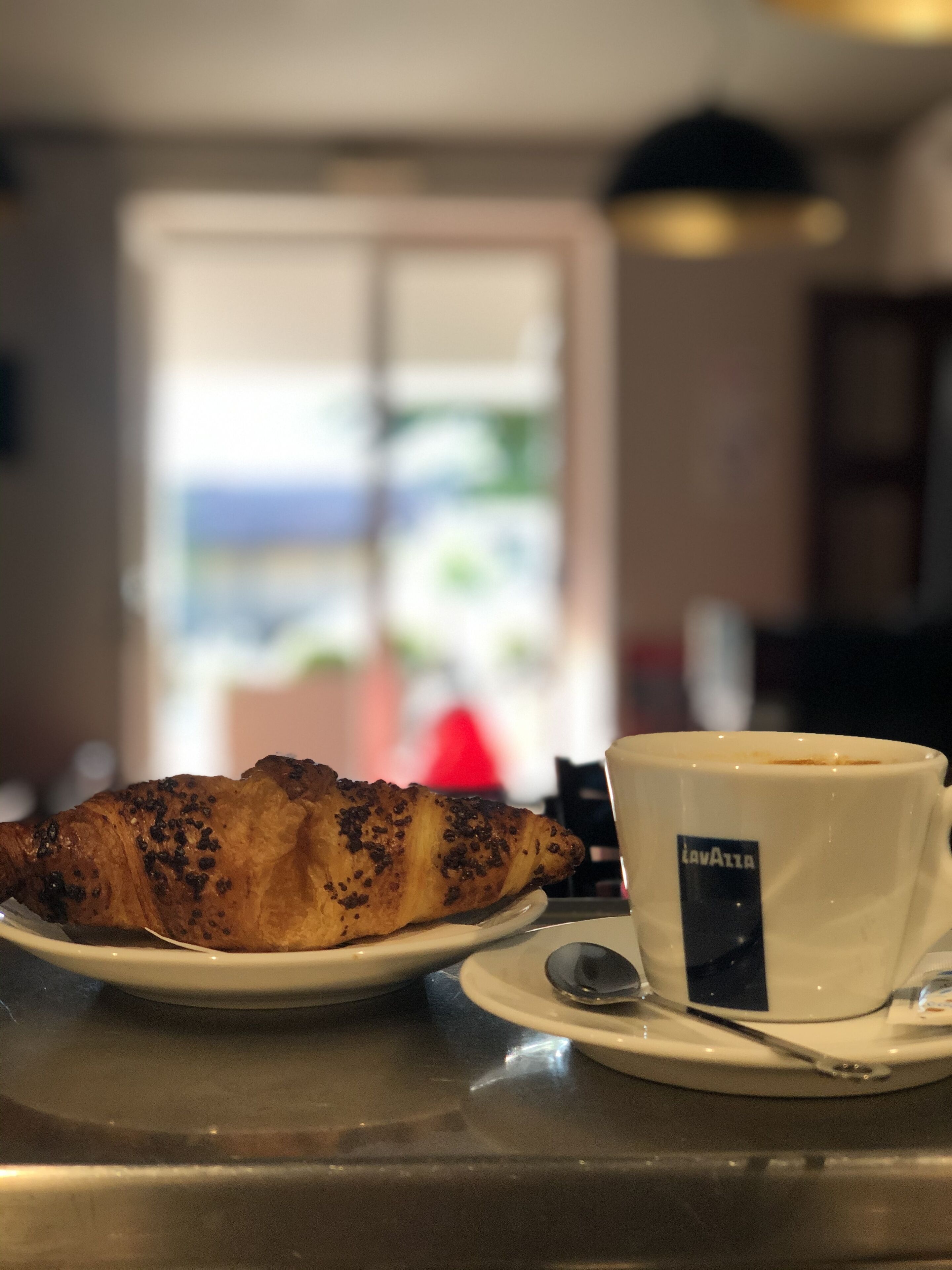 A croissant on a plate next to a cup with a spoon, on a table with a metal surface.