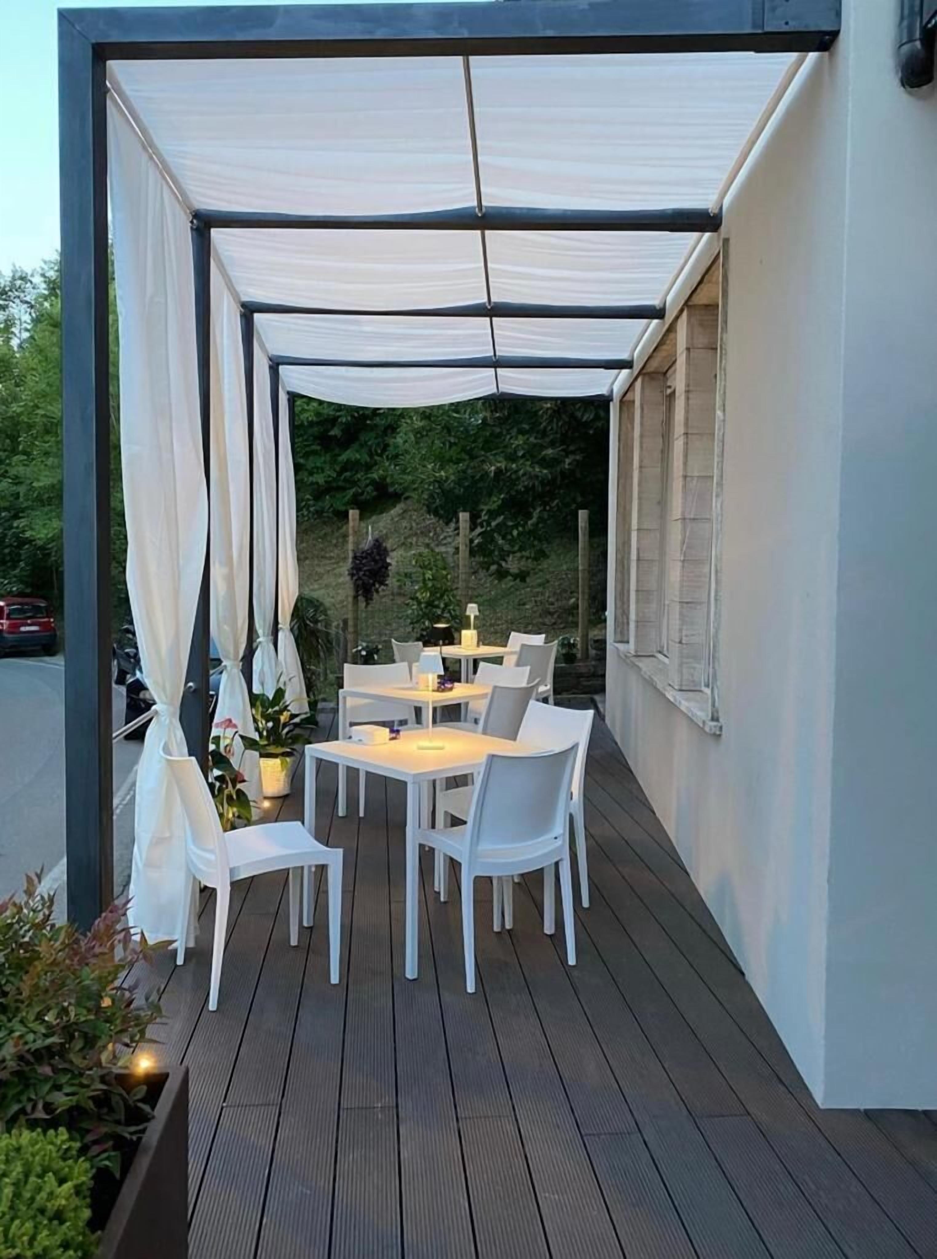 Una veranda esterna con tavoli e sedie bianche, una zona coperta con tende bianche e vista sullo sfondo di vegetazione.