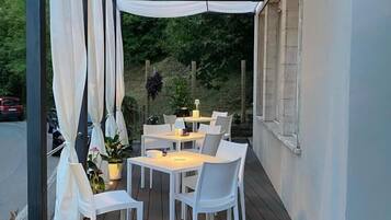 Una veranda esterna con tavoli e sedie bianche, una zona coperta con tende bianche e vista sullo sfondo di vegetazione.