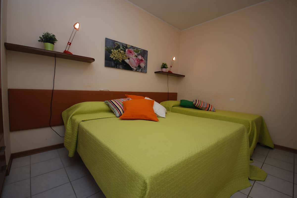 Une chambre avec deux lits recouverts de couvre-lits verts, un tableau au mur et deux petites étagères ornées de plantes et d’une lampe.