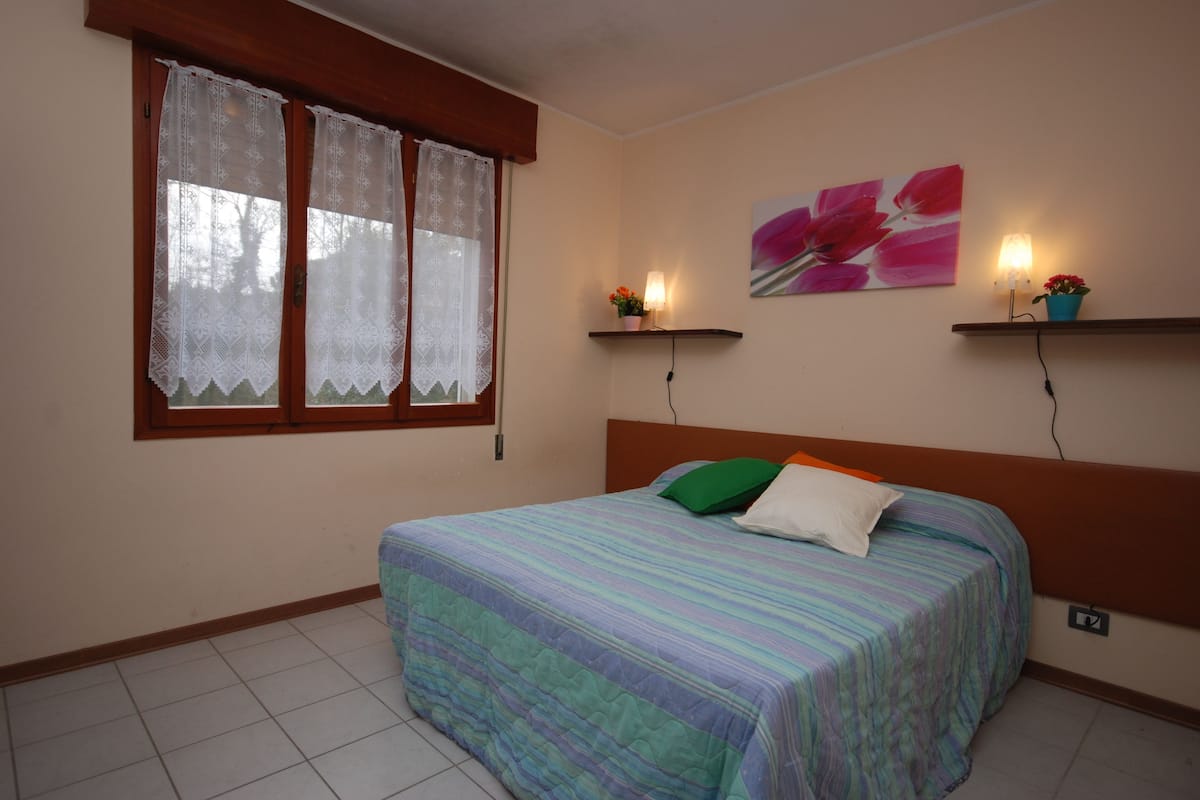 Une chambre à coucher avec un lit, des rideaux en dentelle, un tableau représentant des fleurs et deux étagères fixées au mur, équipées de lumières.
