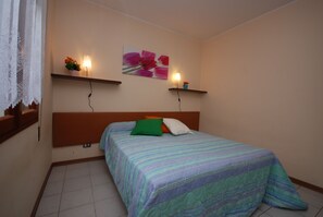 A bedroom with a bed, a headboard, a wall-mounted shelf with a flower, and a wall-mounted light. - Villa Tiziano (Lignano Sabbiadoro)