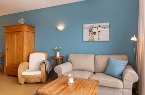 Ein Wohnzimmer mit blauer Wand, einem beigen Sofa, einem Korbstuhl, einem Holzschrank, einem kleinen Tisch mit einer Vase und einem Gemälde mit Strandmotiv.