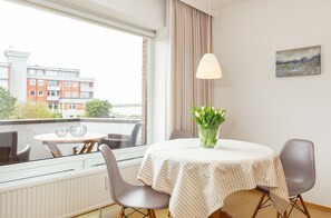 Une pièce avec une table ronde, deux chaises, un vase de fleurs, une lampe suspendue et une fenêtre avec des rideaux.