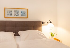 Ein ordentlich bezogenes Bett mit weißen Laken, ein braunes Kopfteil, ein gerahmtes Bild darüber, eine Nachttischlampe und ein kleiner Blumenstrauß auf dem Nachttisch.