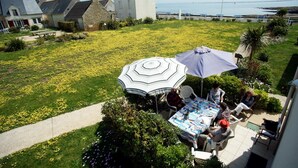A patio area with a table set for dining, surrounded by greenery and a view of the sea. - For rent 2 level house in seaside residence (Le Guilvinec)