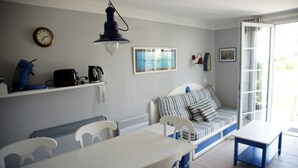 A room with a dining table, chairs, a sofa with striped cushions, a clock, a kettle, a kettle on a shelf, and a window with a view. - For rent 2 level house in seaside residence (Le Guilvinec)