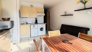 Cocina compacta con lavadora, microondas y armarios de madera. Cuenta con un comedor con mesa y sillas, y una pequeña estantería con un florero.