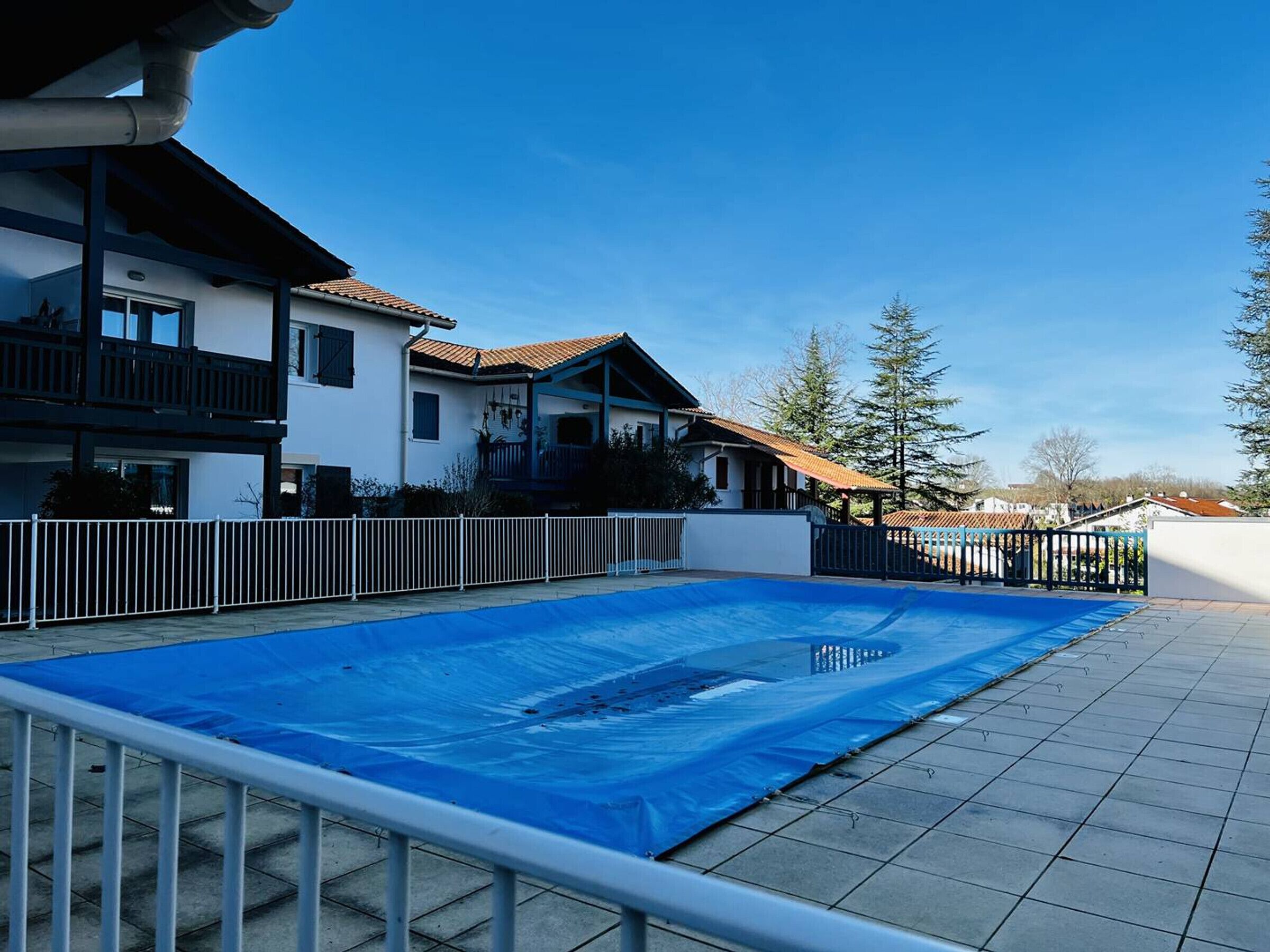Ett bostadsområde med en swimmingpool som är täckt av en blå presenning, omgiven av ett vitt staket, och intilliggande hus med balkonger och tak belagda med tegelpannor.