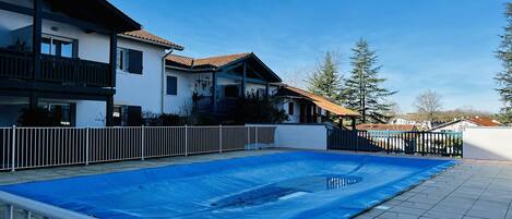 Ett bostadsområde med en swimmingpool som är täckt av en blå presenning, omgiven av ett vitt staket, och intilliggande hus med balkonger och tak belagda med tegelpannor.