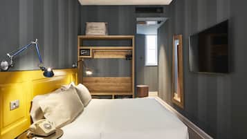 Una camera d'hotel con un letto, una lampada da comodino, una mensola in legno con bottiglie, una televisione e una finestra.