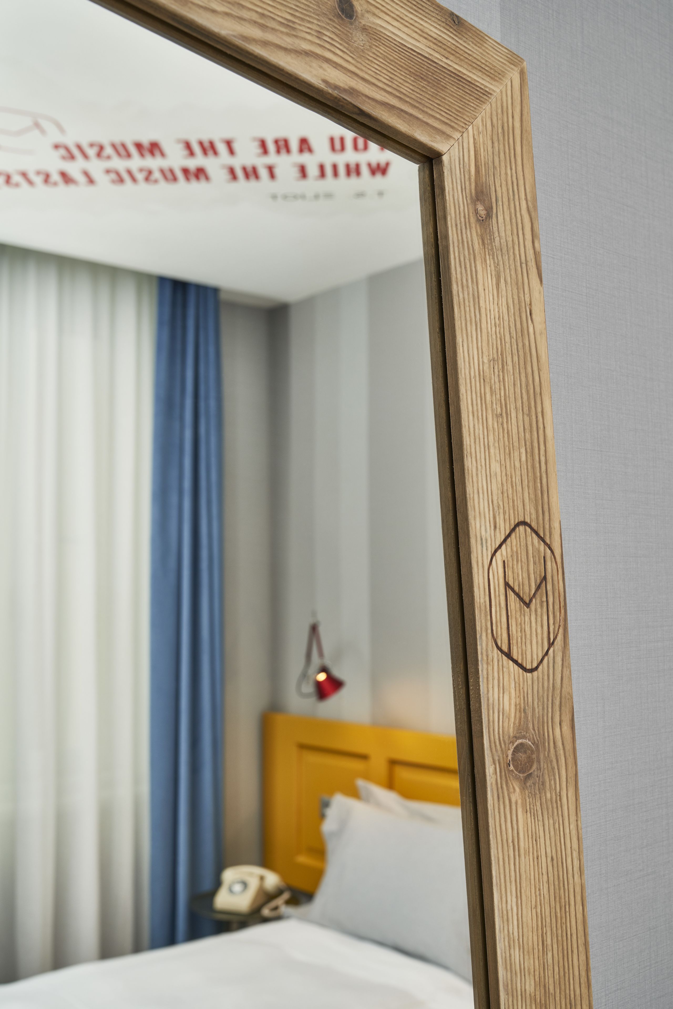 A wooden-framed mirror reflecting a room with a bed, curtains, and a nightstand with a lamp.