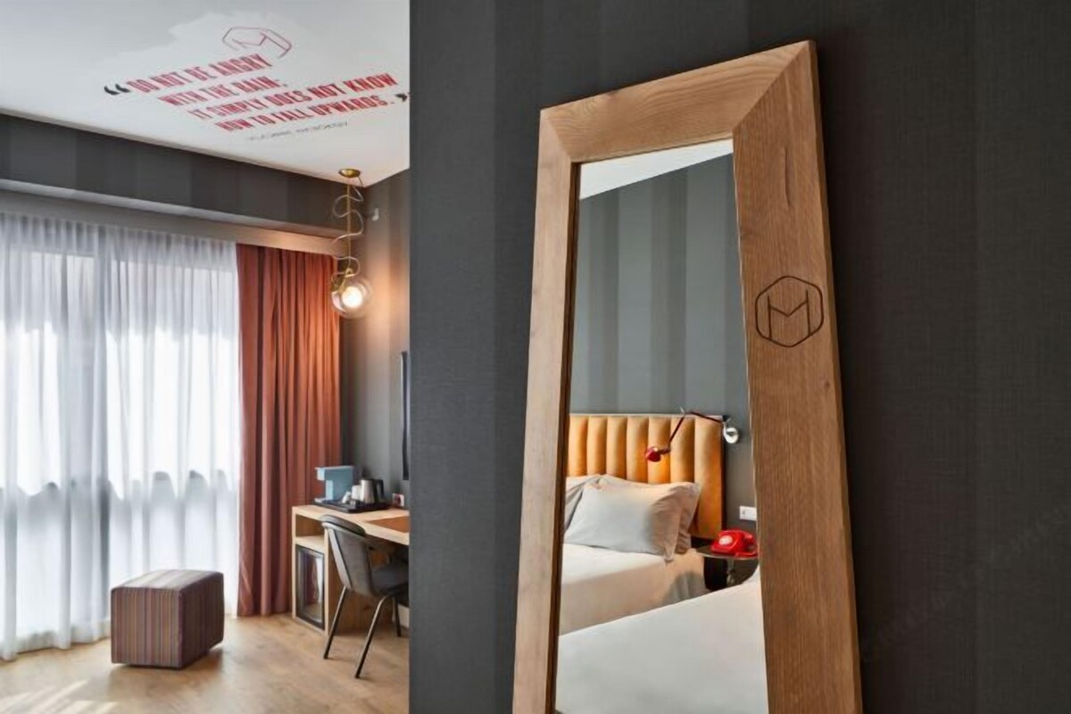 A modern hotel room with a wooden-framed mirror, a desk with a chair, a bed, and a small table with a lamp.