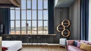 A modern hotel room with a large window, a bed, a desk with two chairs, a sofa, and a small table.