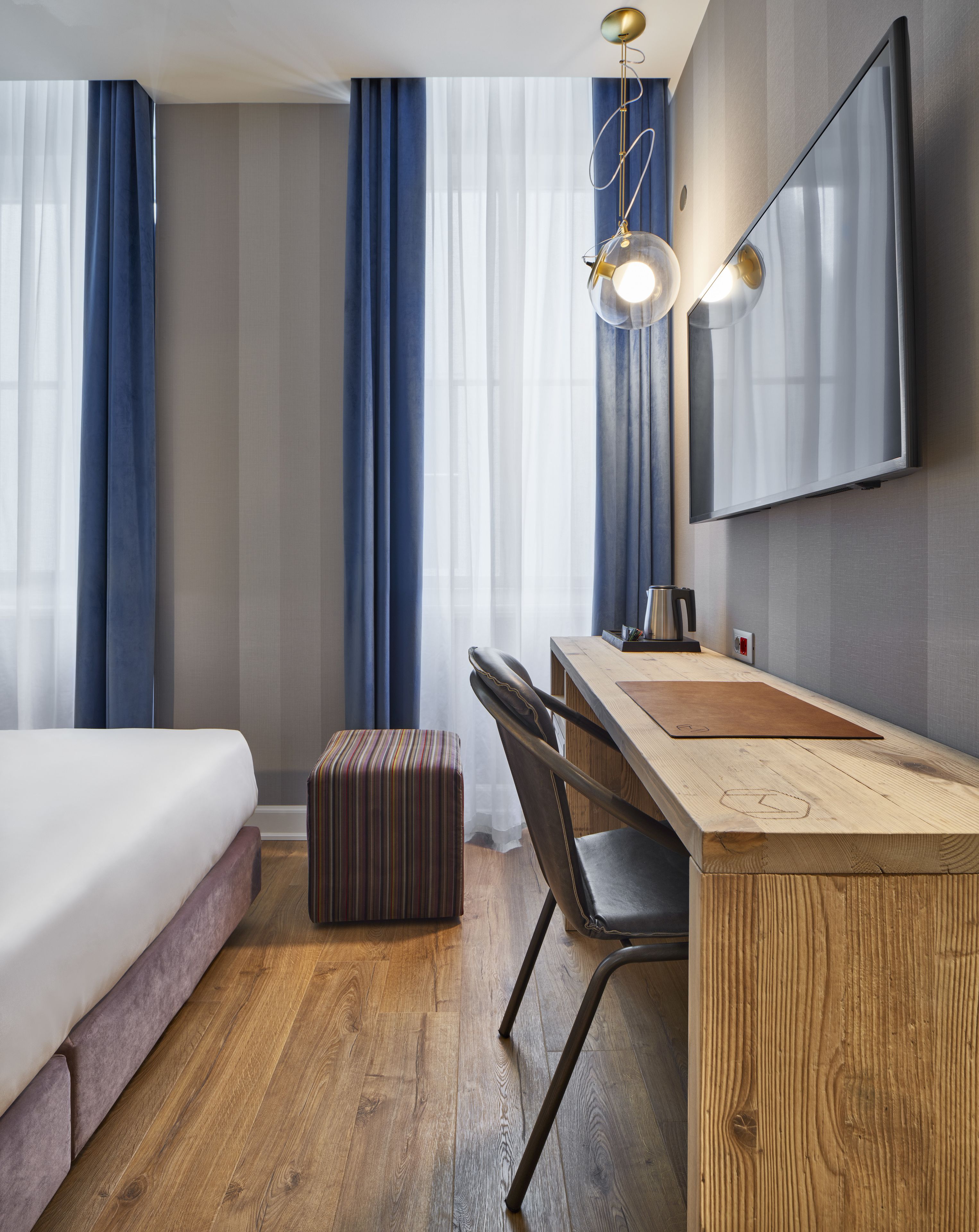 A hotel room with a bed, a wooden desk, a chair, a striped ottoman, a mirror, and a TV.