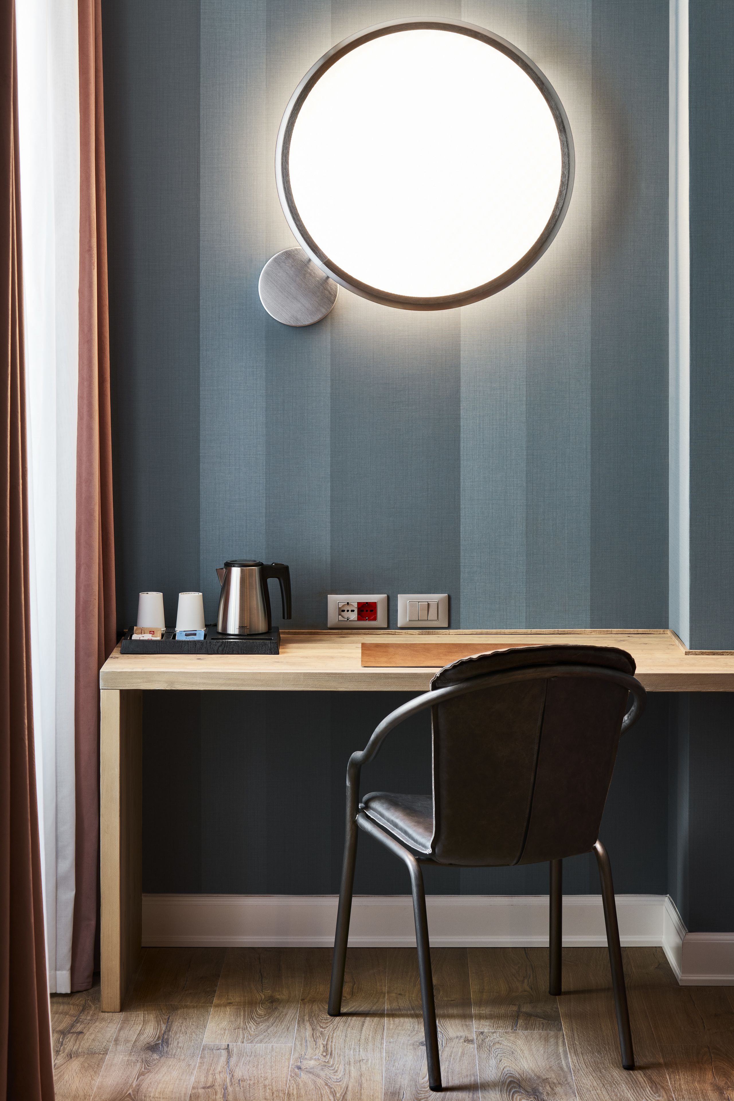 A modern room with a round mirror, a wooden desk, a black chair, and a coffee maker.
