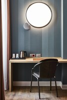 A modern room with a round mirror, a wooden desk, a black chair, and a coffee maker.