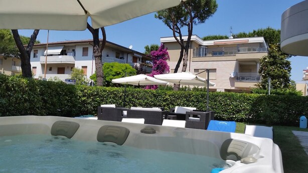 Area con idromassaggio, posti a sedere all'aperto e ombrelloni, circondata da siepi e con edifici sullo sfondo.