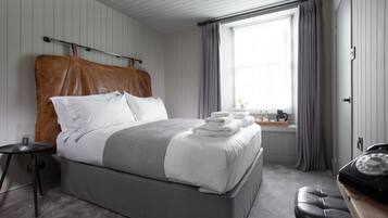 A neatly made bed with white linens and a grey blanket, a bedside table with a lamp, and a window with curtains.