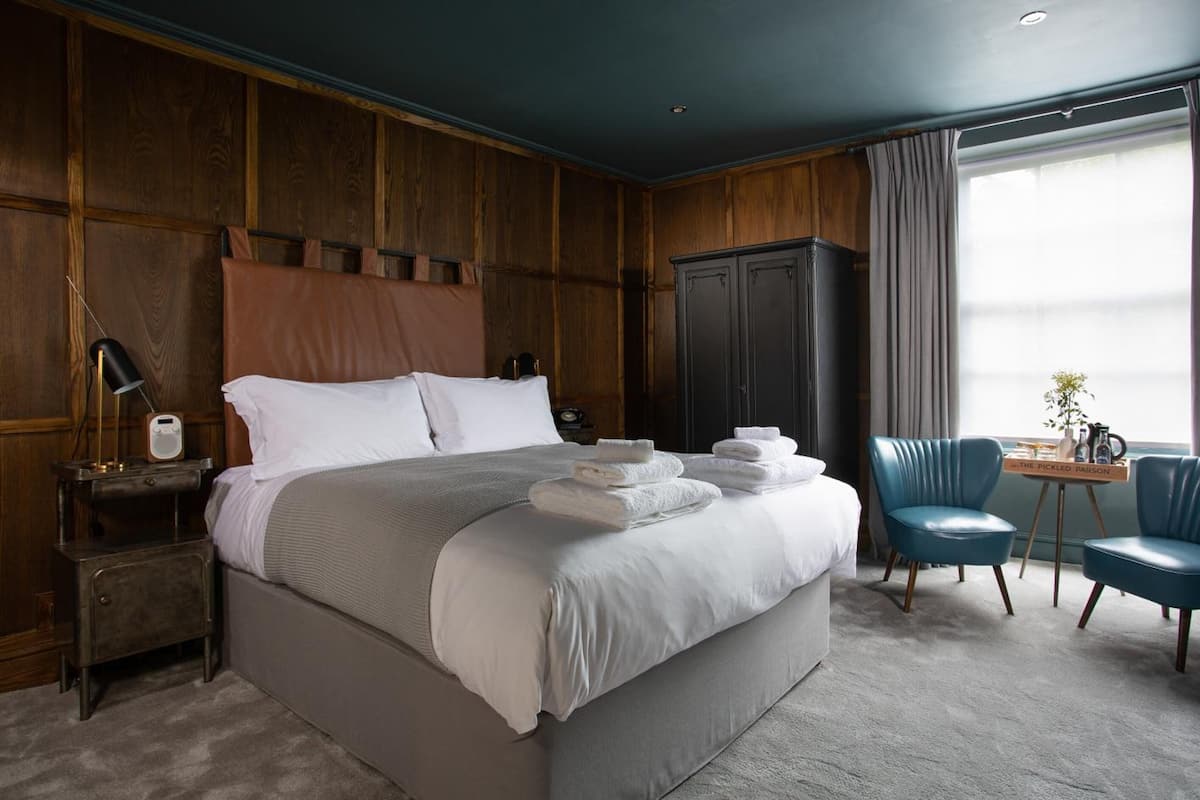 A hotel room with a large bed, a bedside table with a lamp and radio, a wardrobe, a seating area with a chair and table, and a window with curtains.