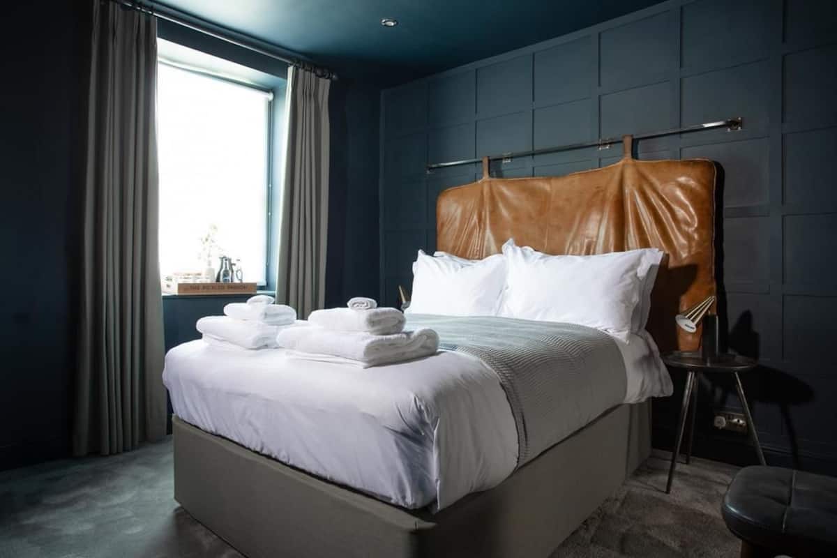 A neatly made bed with white linens and a brown headboard in a room with dark blue walls and a window with curtains.