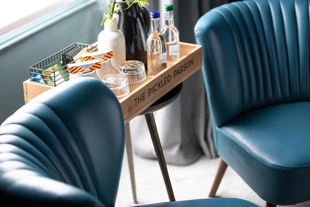 Ein Holztablett mit Flaschen und Gläsern auf einem Tisch, platziert zwischen zwei blauen Stühlen.