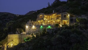 A large, illuminated stone building with multiple levels and balconies, surrounded by dense vegetation and hills. - Felini Palace (Malevizi)