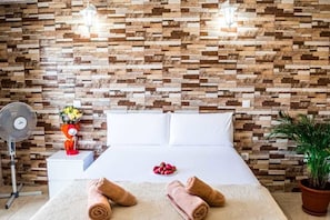 Habitación de hotel con cama, ropa blanca, ventilador, florero con flores y planta en maceta.