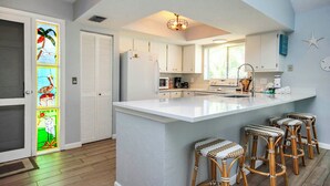 A modern kitchen with white cabinets, a large island with bar stools, and a stained glass door with tropical birds. - On a Whim Captiva Island Beach Cottage (Captiva)