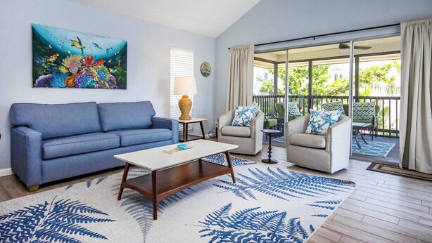 A living room with a blue sofa, a coffee table, and two armchairs with patterned cushions. There's a painting of fish on the wall and a large window with curtains. - On a Whim Captiva Island Beach Cottage (Captiva)
