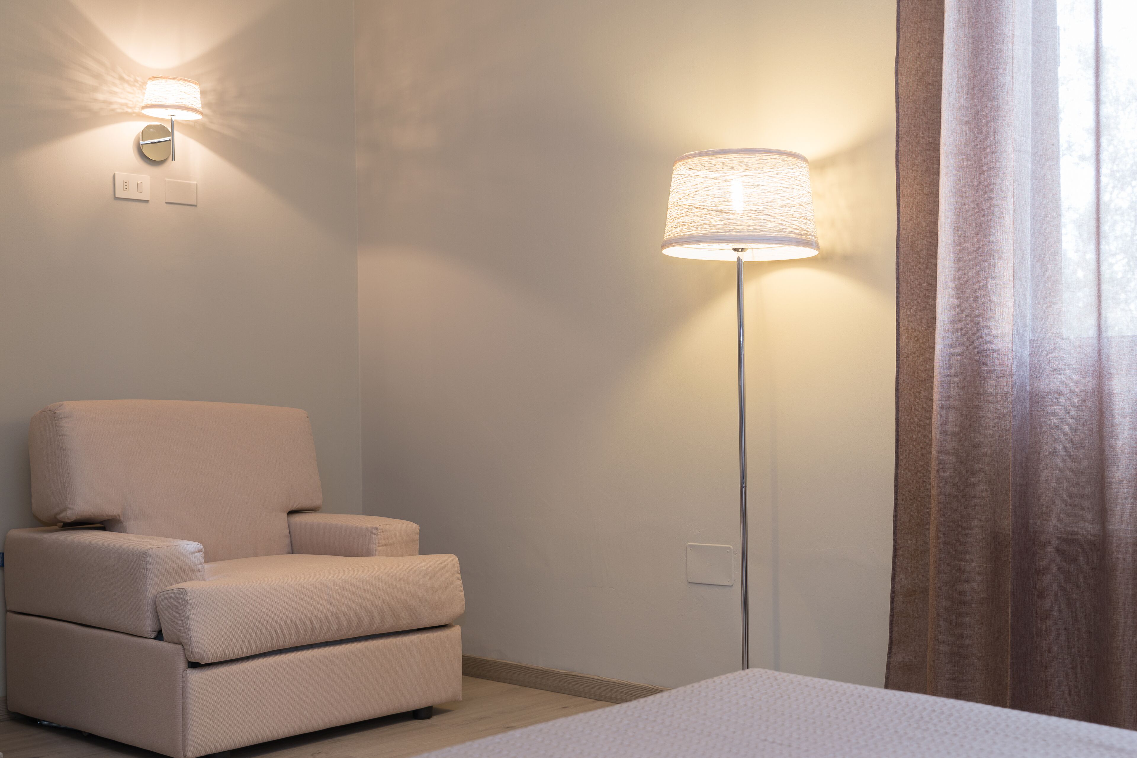 A room with a beige armchair, a floor lamp, and a bed.