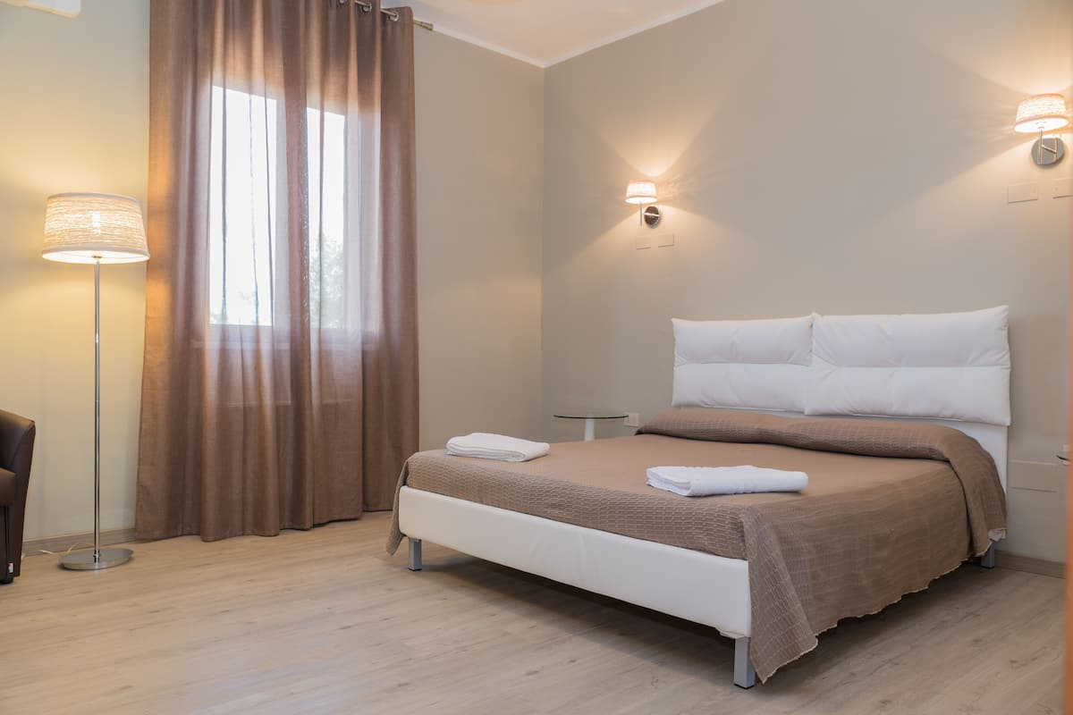 Ein ordentlich bezogenes Bett mit weißer und brauner Bettwäsche, eine Stehlampe und eine Wandleuchte in einem modernen Zimmer mit hellem Holzboden.
