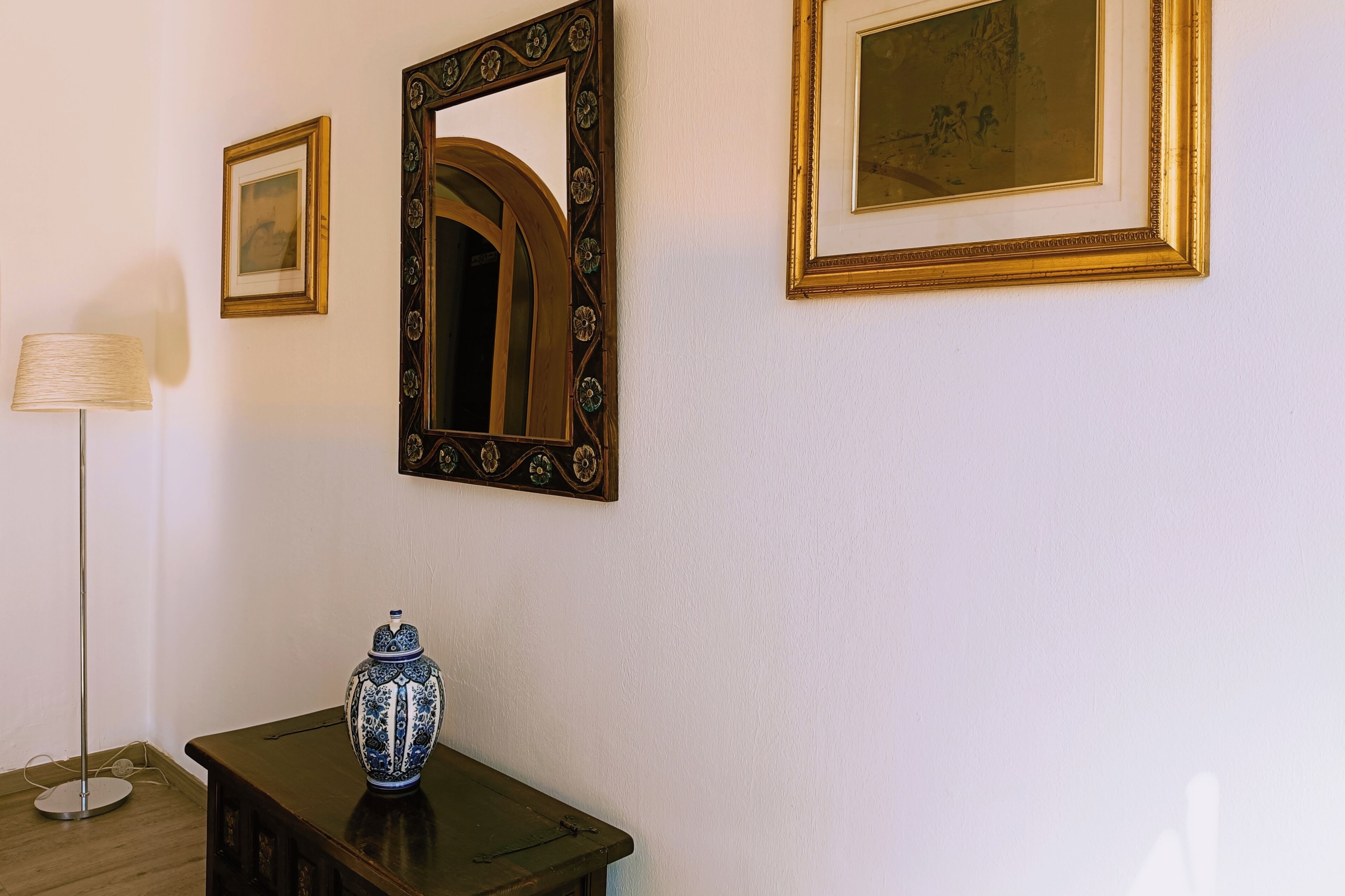 Una stanza con uno specchio, opere d'arte incorniciate, una lampada e un vaso decorativo su un tavolo.