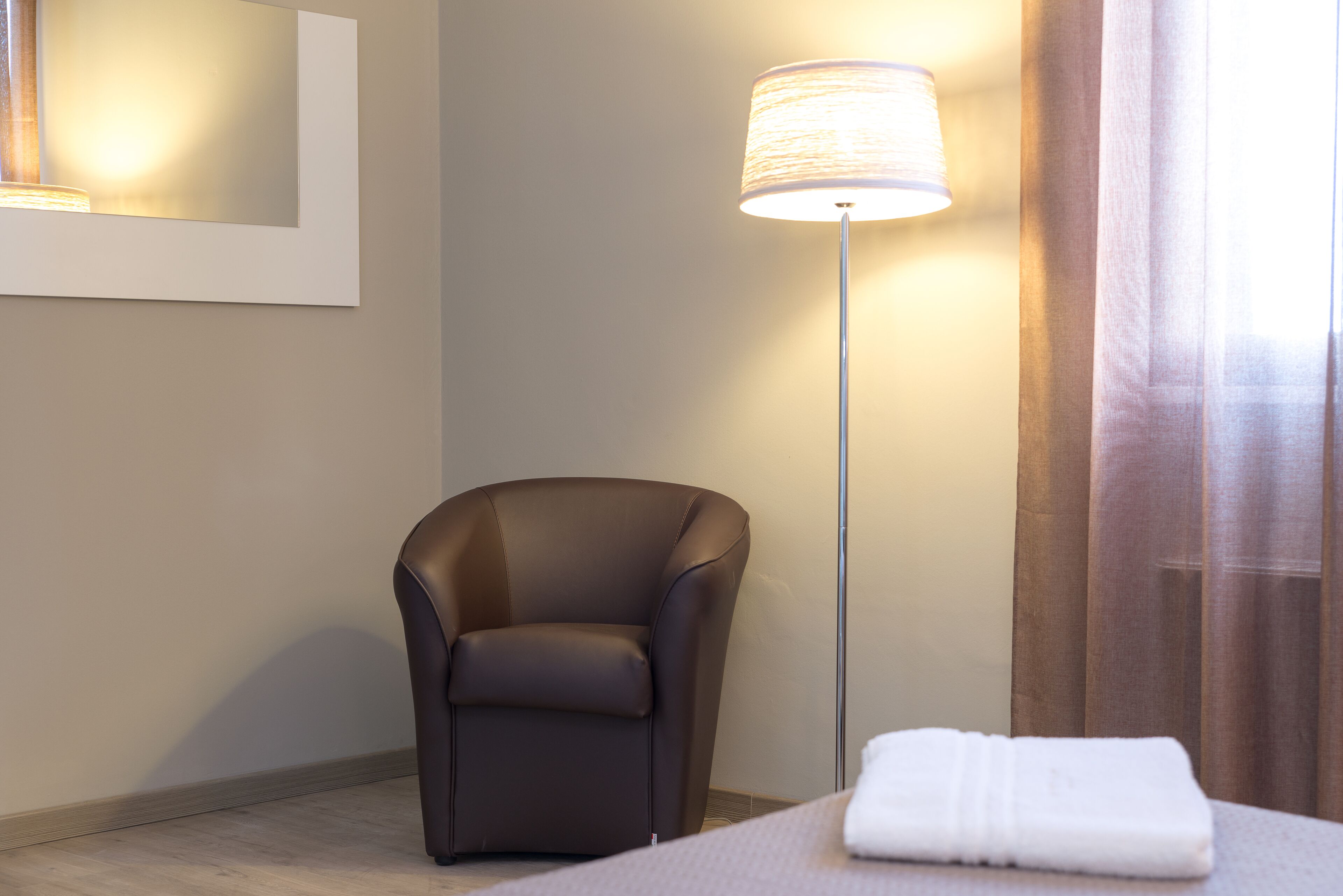 Habitación de hotel moderna con un sillón de cuero oscuro, una lámpara de pie y una toalla blanca doblada con cuidado sobre la cama.