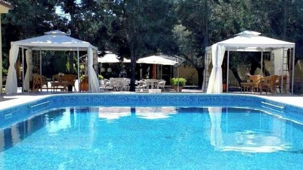 Ein Schwimmbecken mit zwei überdachten Sitzbereichen und Tischen, umgeben von Bäumen.