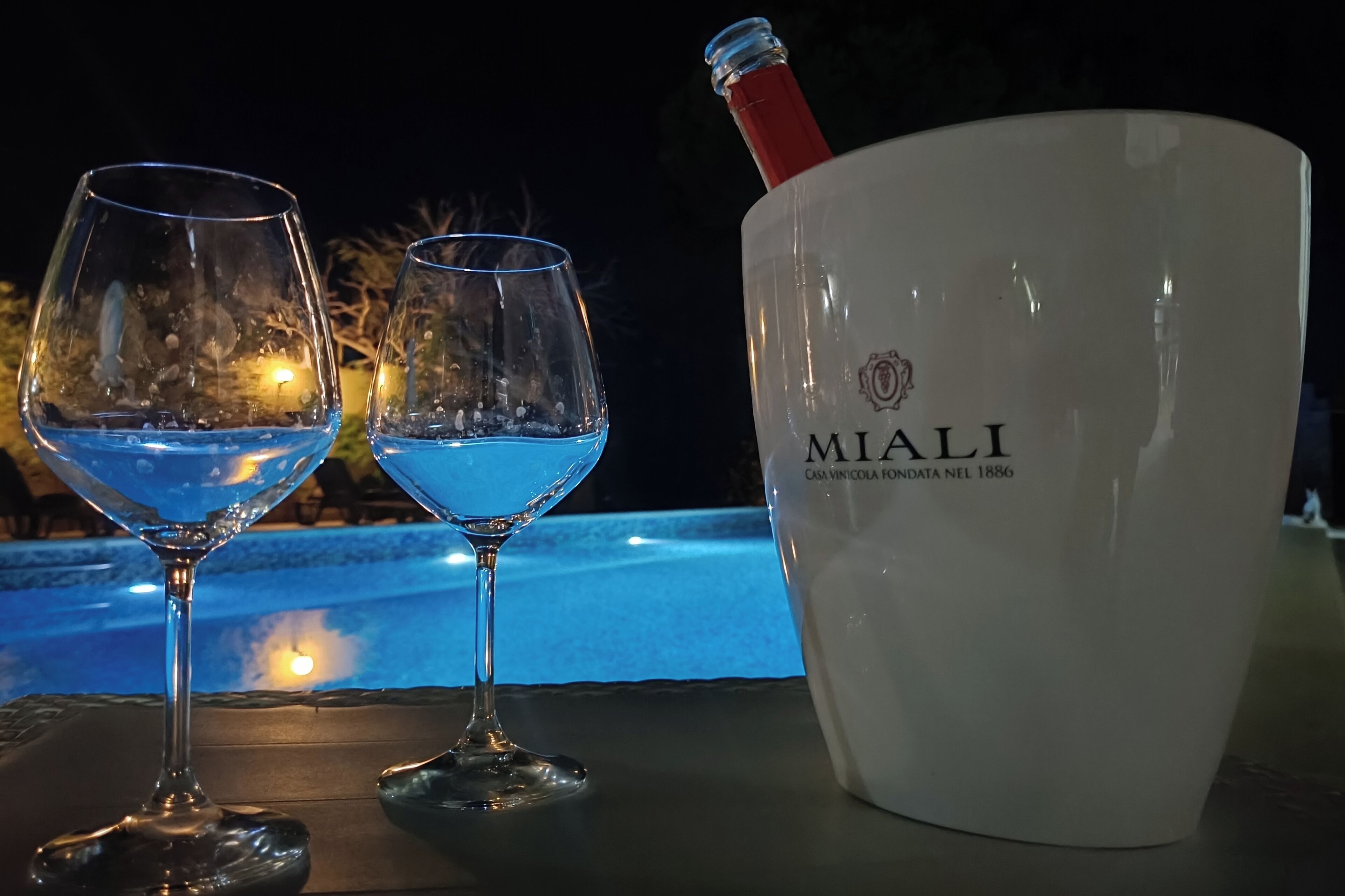 Twee glazen blauwe vloeistof, een fles en een emmer met de tekst 'MIALI' erop, op een tafel, met een zwembad op de achtergrond.