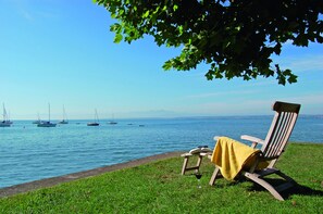 A chair with a yellow towel, a grassy area, and a body of water with boats. - Der Loewen (Hagnau)