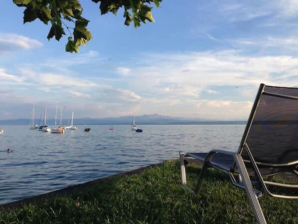 A lakeside view with a chair by the water's edge, boats in the distance, and a mountain range in the background. - Der Loewen (Hagnau)