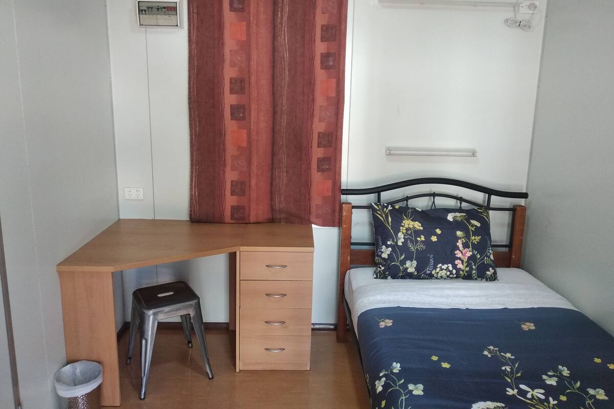 A small, neatly arranged room with a wooden desk, a single bed with floral bedding, a red curtain, and a wall-mounted air conditioner.